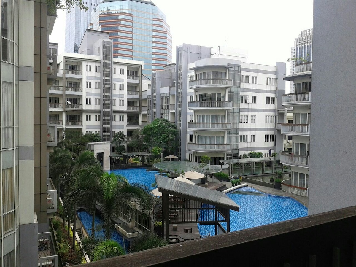 A modern residential complex is showcased, with multiple buildings surrounding a large swimming pool. Lush greenery and palm trees are visible around the pool area, which features shaded seating options. The scene is set against a backdrop of high-rise buildings in the distance.