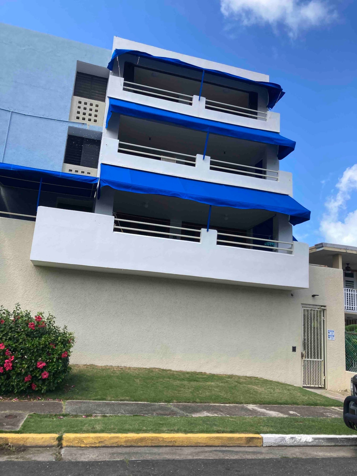 A three-story building showcases a light-colored façade with blue awnings over the balconies. Lush greenery and colorful flowering plants are situated at the base, complementing the structure. The entrance is secured with a gate, hinting at the community's inviting atmosphere.
