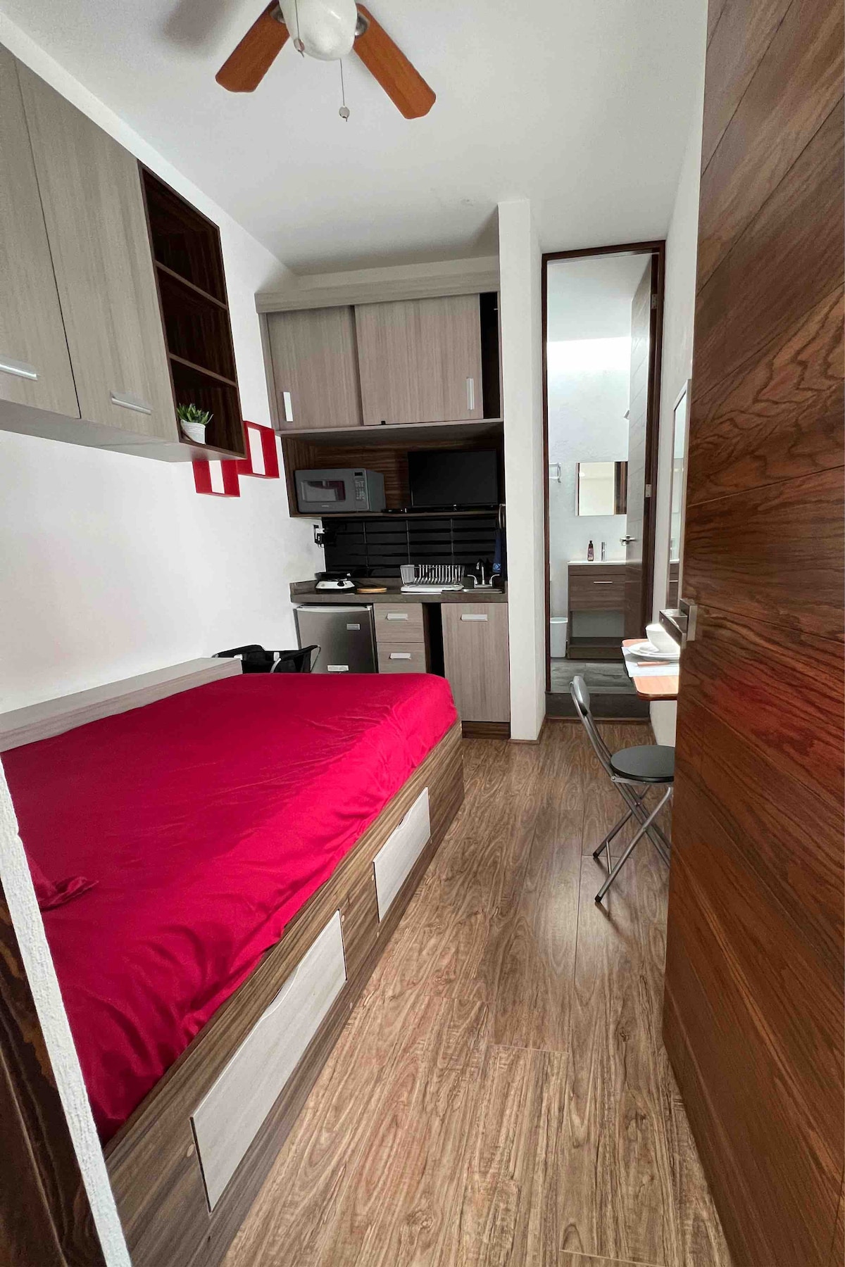 A compact room features a cozy bed covered with a red blanket. Kitchen appliances are arranged in a small kitchenette, with storage cabinets above. A workspace with two chairs is positioned beside a mirror, while wooden flooring extends throughout the space.