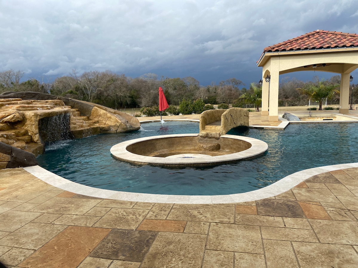 The outdoor pool area features a circular, elevated spa surrounded by a soothing waterfall. A bright red umbrella adds a pop of color beside the pool, which is framed by stone accents and lush landscaping under a dramatic sky.
