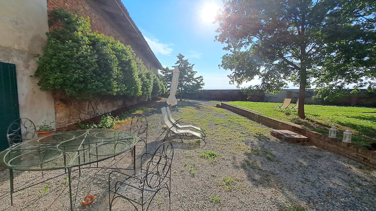 An outdoor garden area features a gravel surface with several lounge chairs positioned for relaxation. A circular glass table is surrounded by four metal chairs. Lush greenery and tall trees provide shade, with sunshine filtering through, creating a serene atmosphere.