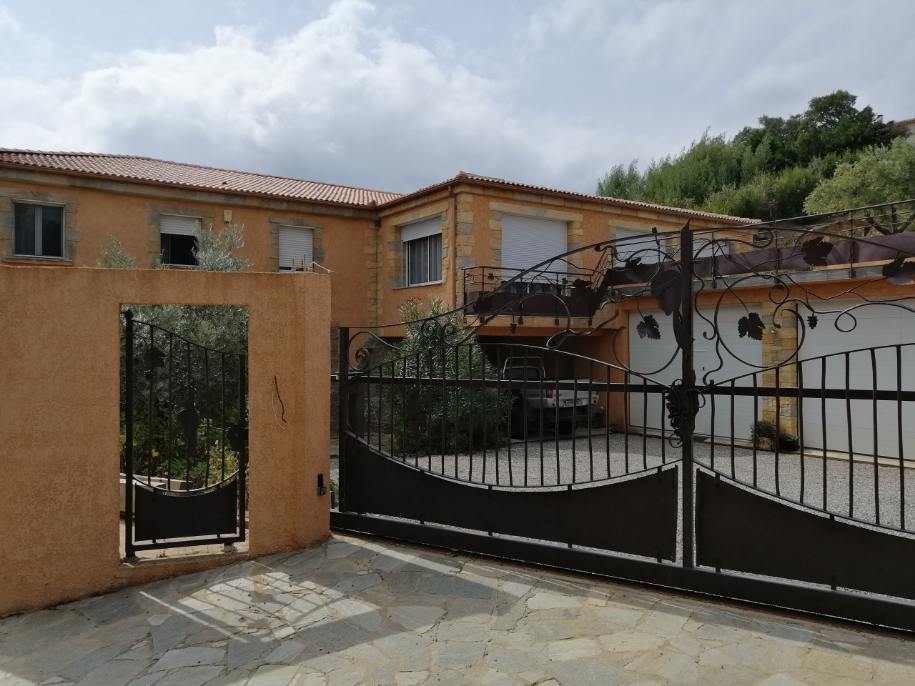 A gated entrance leads to a spacious property with a stone facade and a tiled roof. Overhead, clouds are visible, hinting at an inviting outdoor space. The driveway allows for accessible parking, with vehicles observed near the structure, surrounded by greenery.
