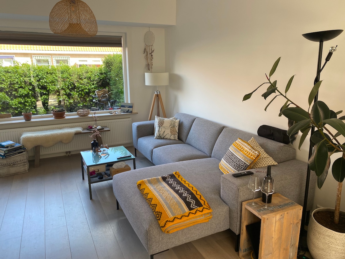 A spacious living area is highlighted by a light grey sectional sofa adorned with vibrant throw pillows and a cozy blanket. Large windows provide natural light, offering views of lush greenery outside. A minimalist coffee table and modern floor lamp complement the room's relaxed ambiance.