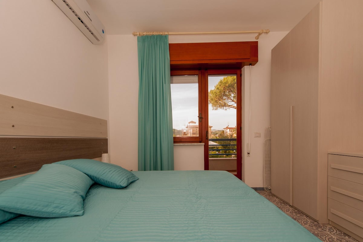 A bright bedroom features a spacious bed dressed in aqua blue linens, complemented by a light curtain framing an inviting window. Natural light streams in, highlighting the wood and neutral tones of the room. A compact wardrobe is positioned nearby, with a view of trees outside.