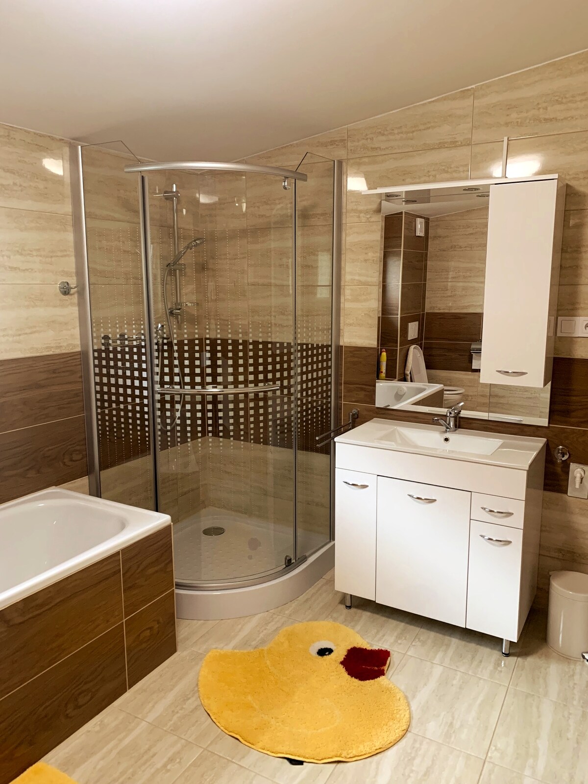 A modern bathroom features a walk-in shower enclosed by glass, alongside a freestanding bathtub. A white vanity unit with storage is positioned beside a large mirror, and a soft bath mat is placed on the floor for comfort.