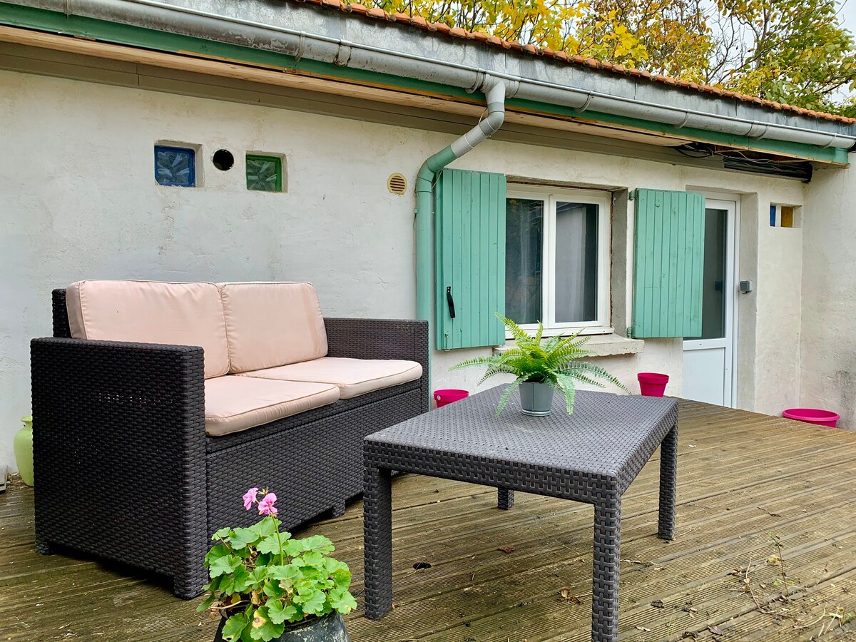 An outdoor terrace is visible, featuring a brown wicker sofa with light cushions and a matching table. Surrounding greenery adds to the tranquil atmosphere. Two light blue shutters frame a small window, and potted plants are positioned around the area, enhancing the inviting outdoor space.