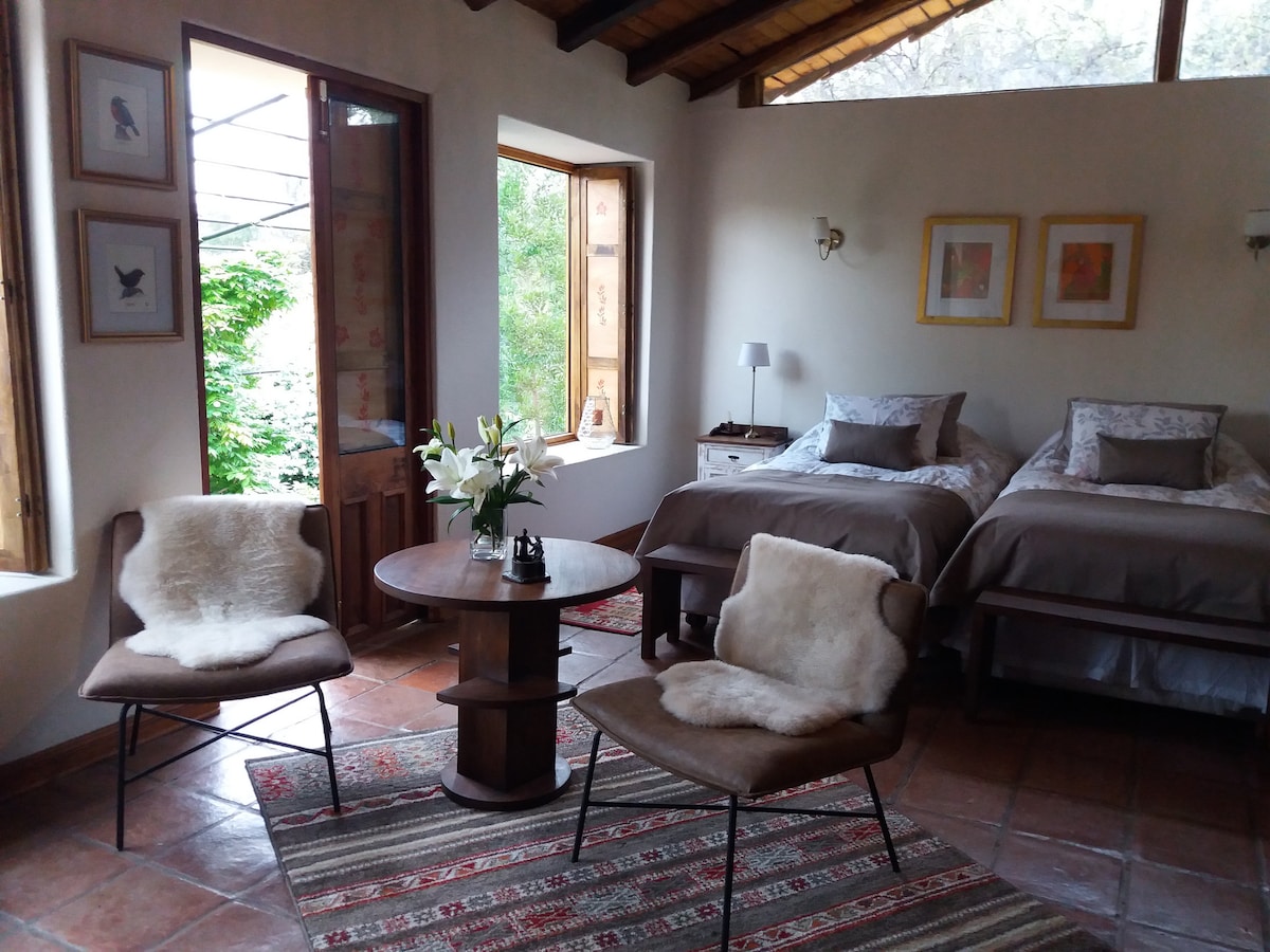 A spacious room includes two beds with neutral bedding, complemented by soft throw pillows. Two cozy chairs, lined with fur, are positioned around a small wooden table. Natural light filters in through large windows, enhancing the room's inviting ambiance.