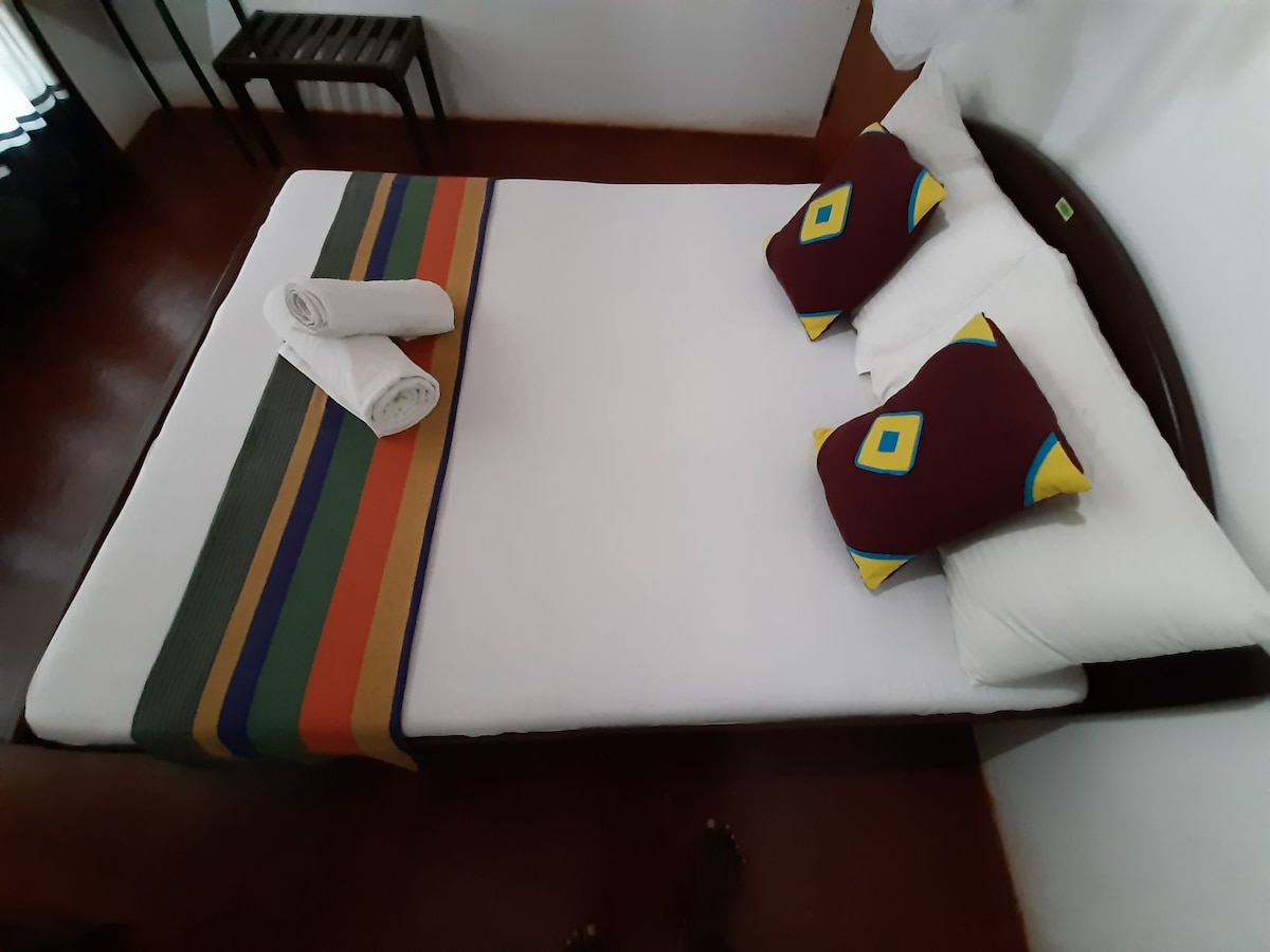 A neatly arranged bed features a white sheet and colorful pillows with geometric patterns. Two rolled towels are placed at the foot of the bed. A small table is positioned against the wall, providing a functional space within the room.