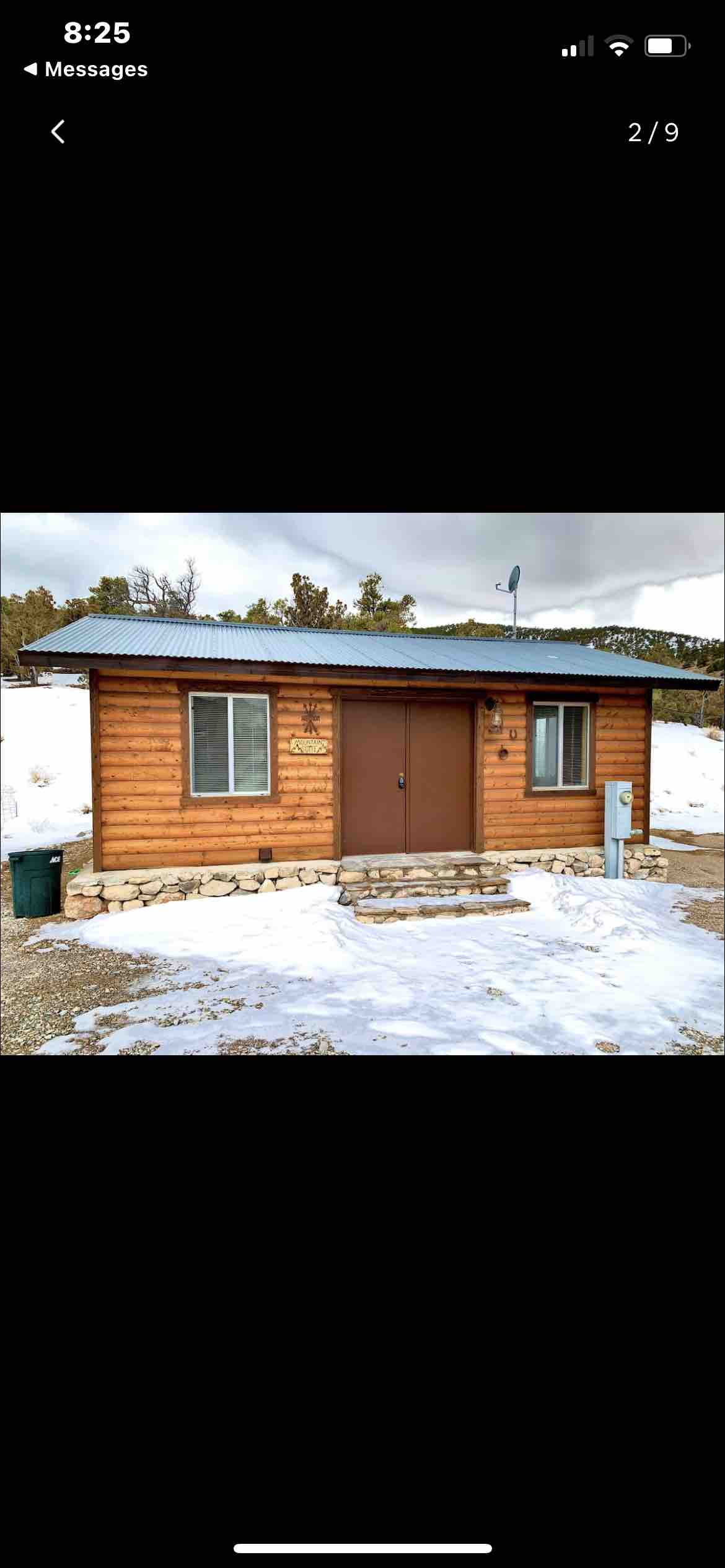 A cozy studio cabin is set against a snowy landscape, showcasing a wooden exterior with a stone foundation. Two windows frame the front door, allowing natural light to brighten the entrance. A satellite dish is mounted on the roof, complementing the serene mountain surroundings.