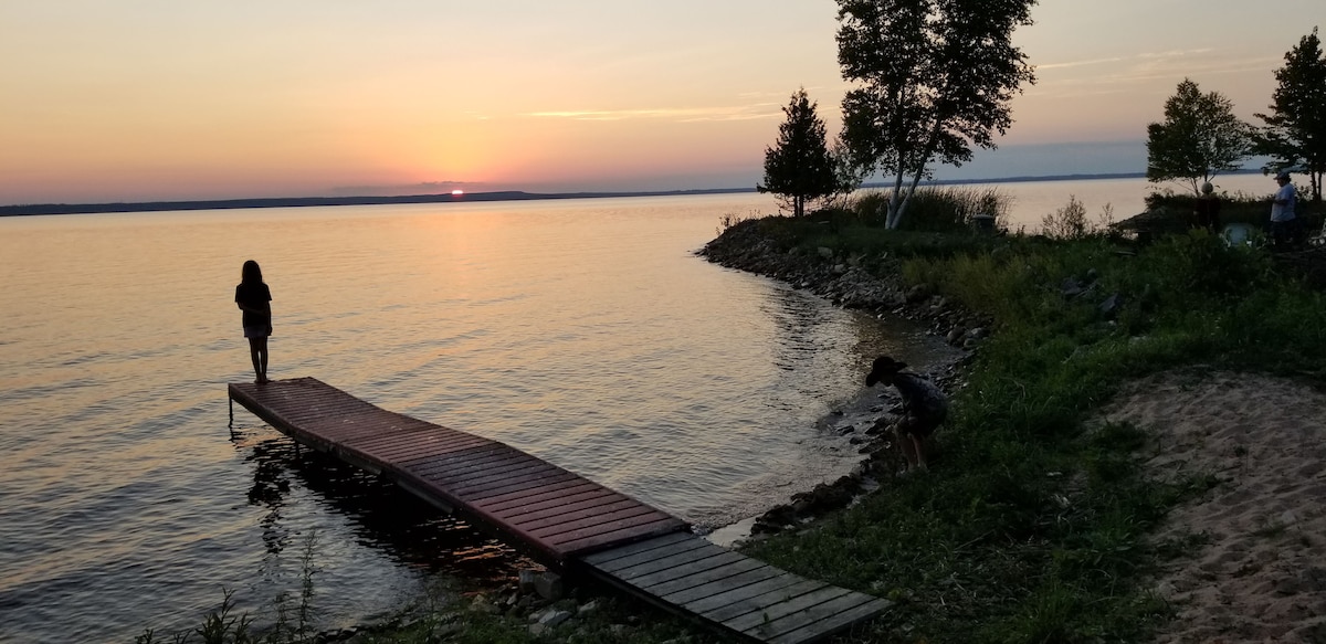 The image captures a serene sunset over Indian Lake, with soft hues reflecting on the water. A wooden dock extends into the lake, while a sandy beach area is visible at the shore. Silhouettes of people are seen enjoying the peaceful surroundings.
