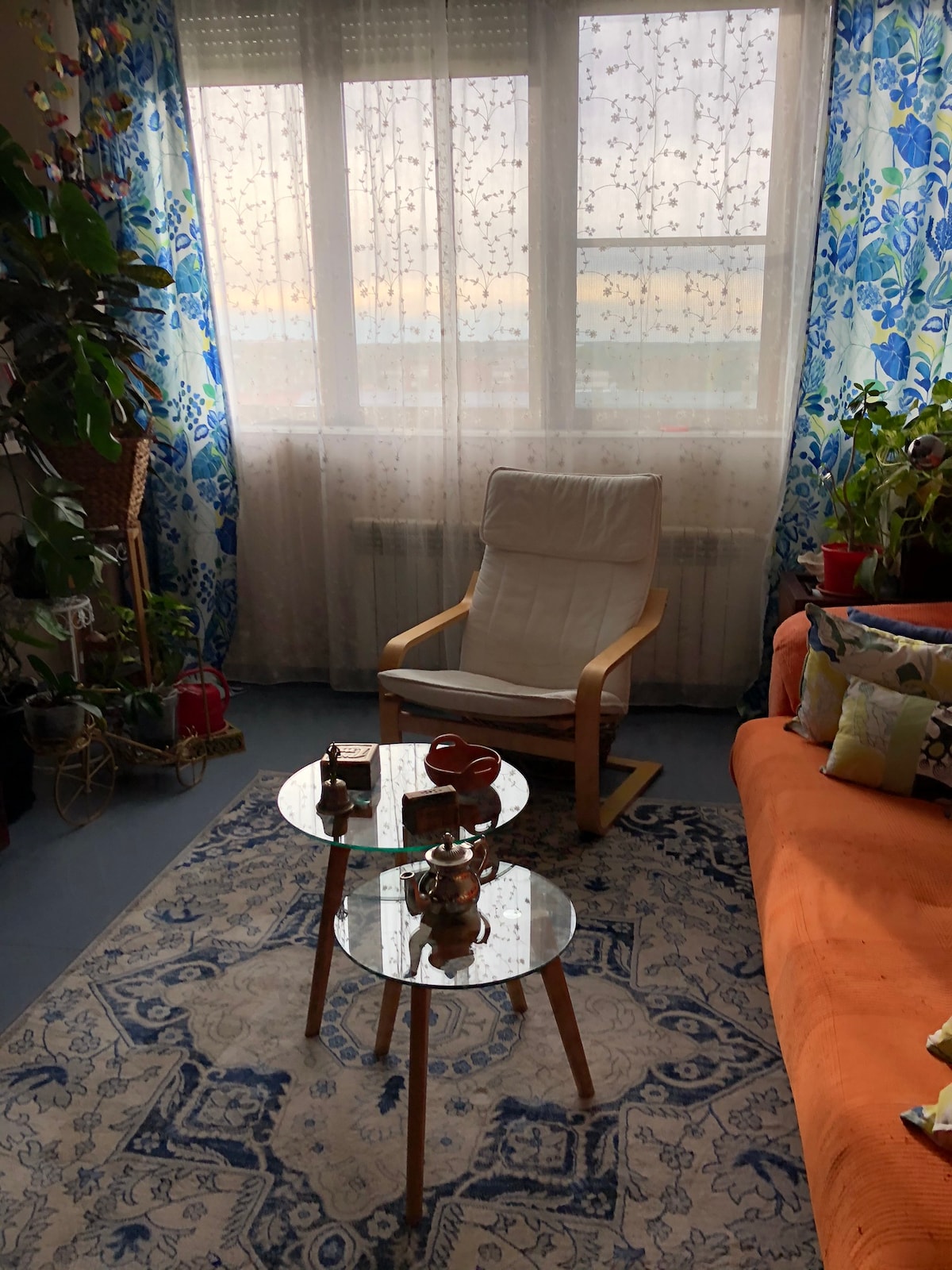 A cozy living area features an orange sofa paired with a white armchair. A glass-top coffee table sits in front, showcasing decorative items. Natural light filters through sheer curtains, illuminating the floral-patterned drapes and creating a warm ambiance. Potted plants add a touch of greenery.