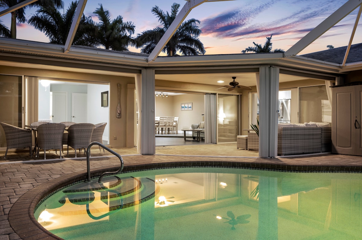 The image captures a heated saltwater pool surrounded by a screened enclosure. Nearby, outdoor seating areas are visible along with a spacious interior that features large glass doors. A serene sunset sky can be seen in the background, complemented by palm trees.