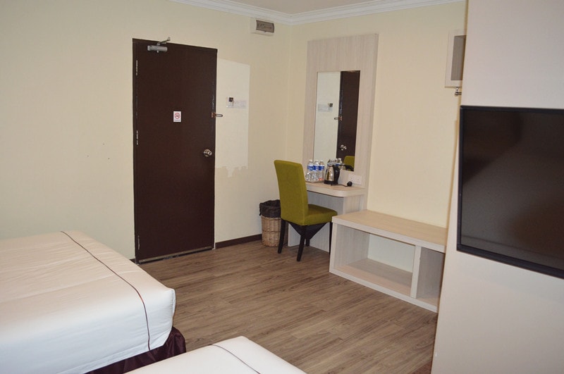 A room features two beds with white linens, wooden flooring, and simple decor. A wooden desk and chair are positioned near a wall mirror, with amenities such as bottled water visible. A dark door and a wall-mounted TV are also present.