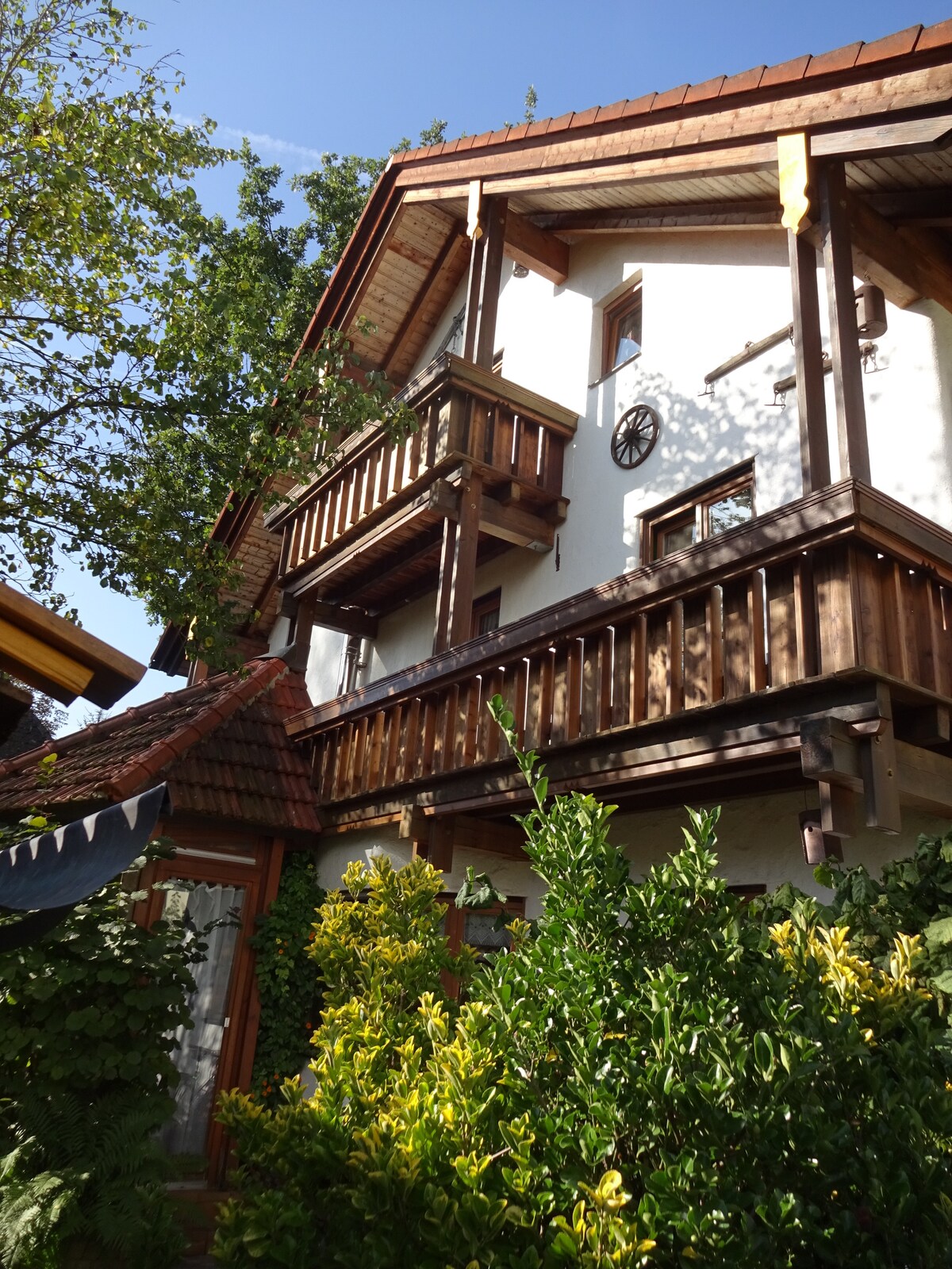 The exterior of a multi-level house is presented, featuring wooden balconies and a combination of traditional textures. Lush greenery surrounds the structure, offering a sense of tranquility. The roof showcases distinctive wooden shingles, complemented by the vibrant colors of the landscaped garden.