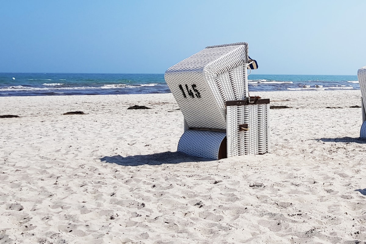 Two woven beach chairs are positioned on a sandy beach, facing the calm sea. The chairs, designed for relaxation, rest beside the gentle waves, creating a serene coastal setting. The clear blue sky above complements the tranquil atmosphere of the beach.
