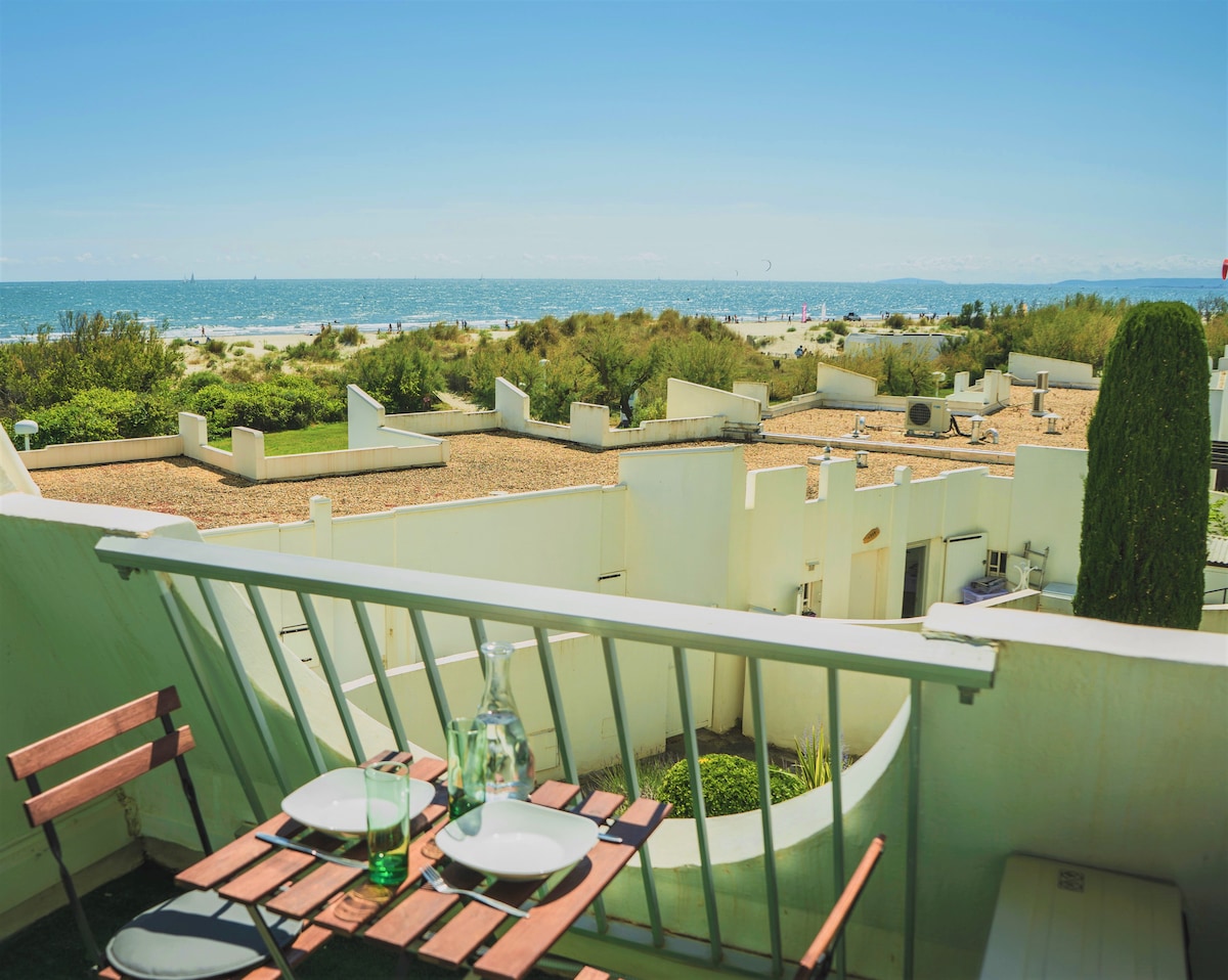 A serene balcony view showcases a dining table set for two, overlooking the sea. The distant shoreline is visible with gentle waves and sandy beach. Lush greenery adds softness to the scene, creating a tranquil setting perfect for enjoying outdoor meals.