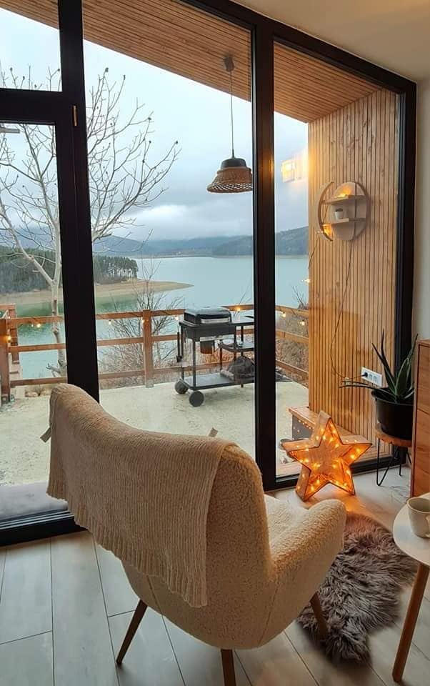 A cozy seating area is positioned near large glass doors, offering a view of the lake. A star-shaped light decoration sits on a soft rug, while an outdoor grill is visible on a private terrace surrounded by natural scenery.