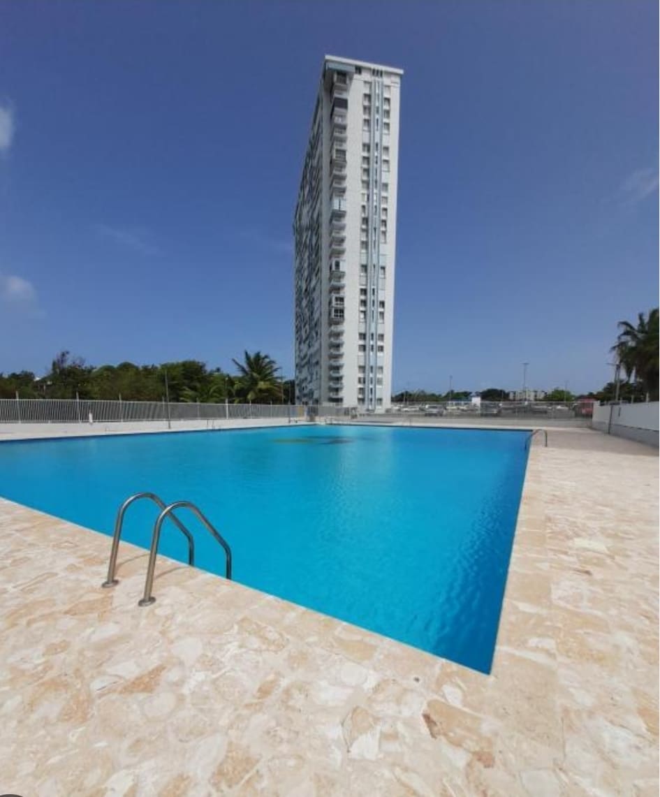 A large swimming pool is framed by a smooth stone deck, with crystal-clear water reflecting the sunlight. In the background, a tall building rises against a clear blue sky, surrounded by palm trees, creating a serene atmosphere.