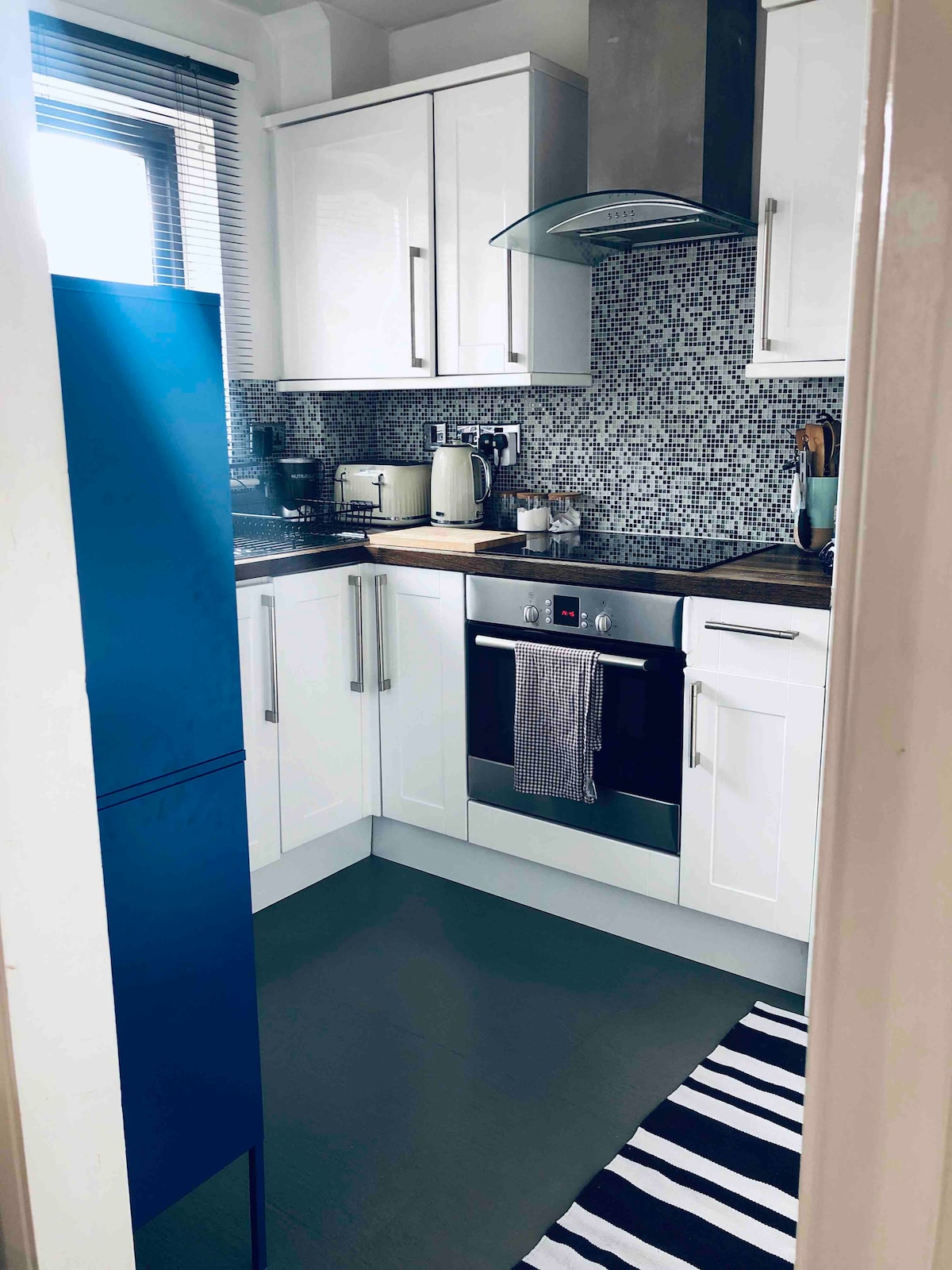 The kitchen features modern cabinetry in white, complemented by a mosaic tile backsplash. A sleek blue fridge stands next to an electric oven and stove. Essential kitchenware is organized on the countertop, with a striped mat placed on the floor for added detail.