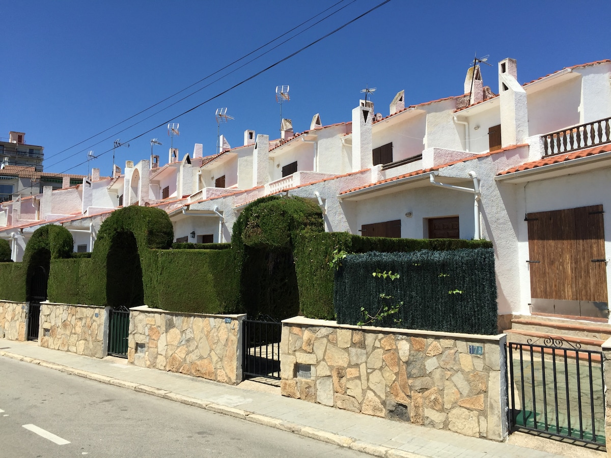A row of white structures can be seen, featuring terracotta roofs and neatly trimmed hedge borders. The scene captures a clear blue sky above, with green plants positioned along the stone boundary wall, providing a fresh and crisp appearance.