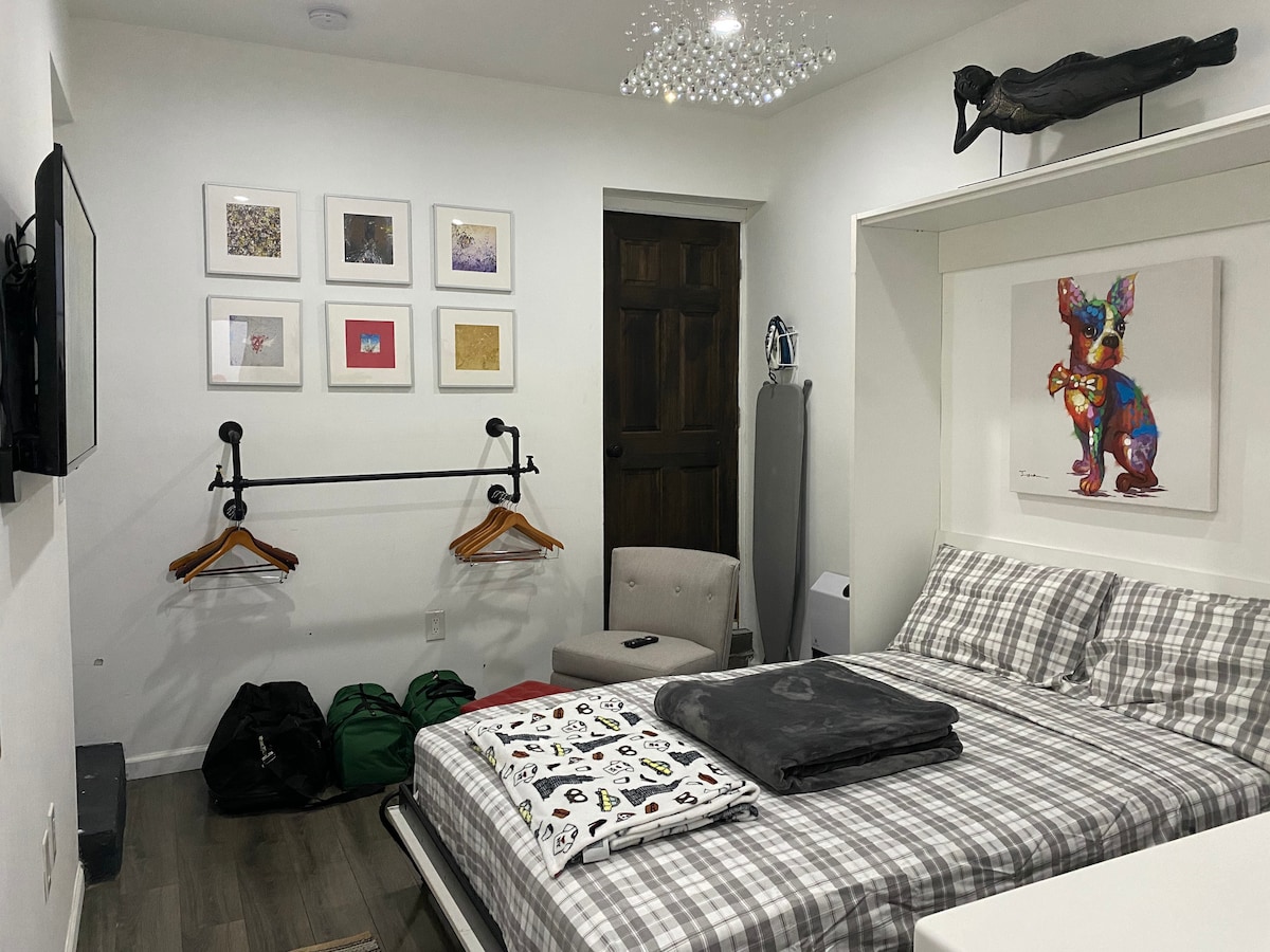A modern studio is presented, featuring a spacious Murphy bed dressed in plaid linens. A colorful artwork and framed prints adorn the walls. An armchair and an ironing board are visible, along with a wall-mounted TV. There are several bags placed at the foot of the bed.