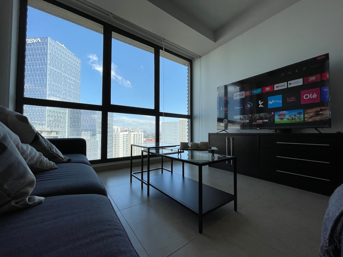 A modern living area is showcased with a large flat-screen TV mounted on a wall unit. A sleek coffee table is positioned in front of a spacious sofa, while large windows provide a view of the skyline, allowing natural light to fill the room.
