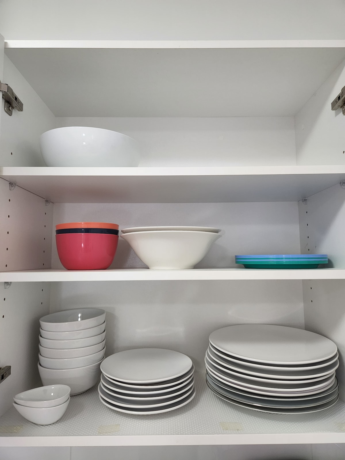 A kitchen cabinet is shown with various dishware neatly arranged. The shelves include a mix of bowls and plates in white, along with a few colorful items, such as a pink bowl and a blue plate. All items appear organized and ready for use.