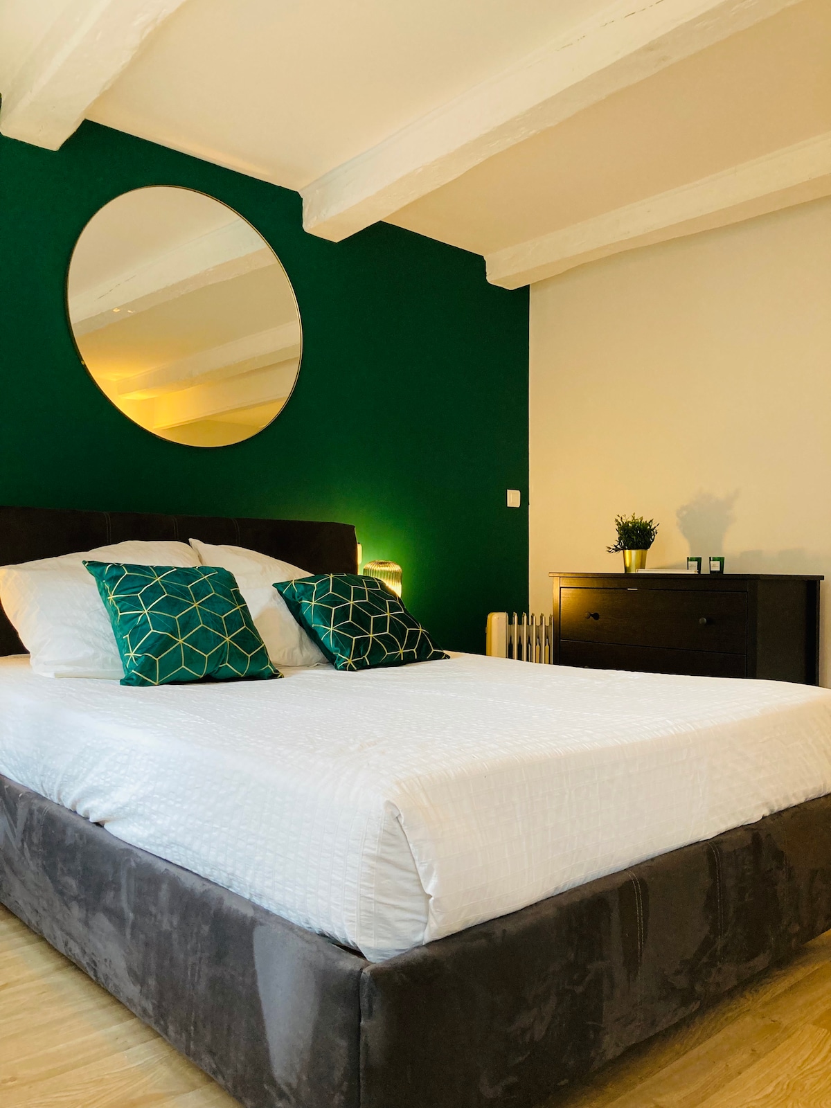 A comfortable sleeping area is highlighted by a spacious bed adorned with decorative cushions. A round mirror reflects the warm light, while a dark wood dresser and a small plant add subtle details to the cozy space.