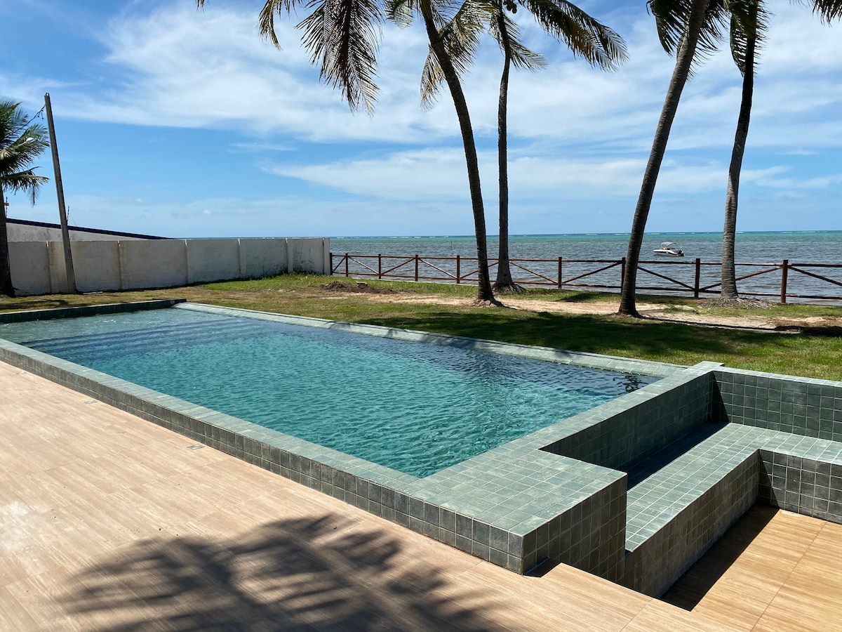 An infinity pool is situated beside the beach, offering views of the ocean. Tall palm trees frame the area, and a spacious deck leads to the water. The pool's tiled surface reflects sunlight, enhancing the tranquil setting.