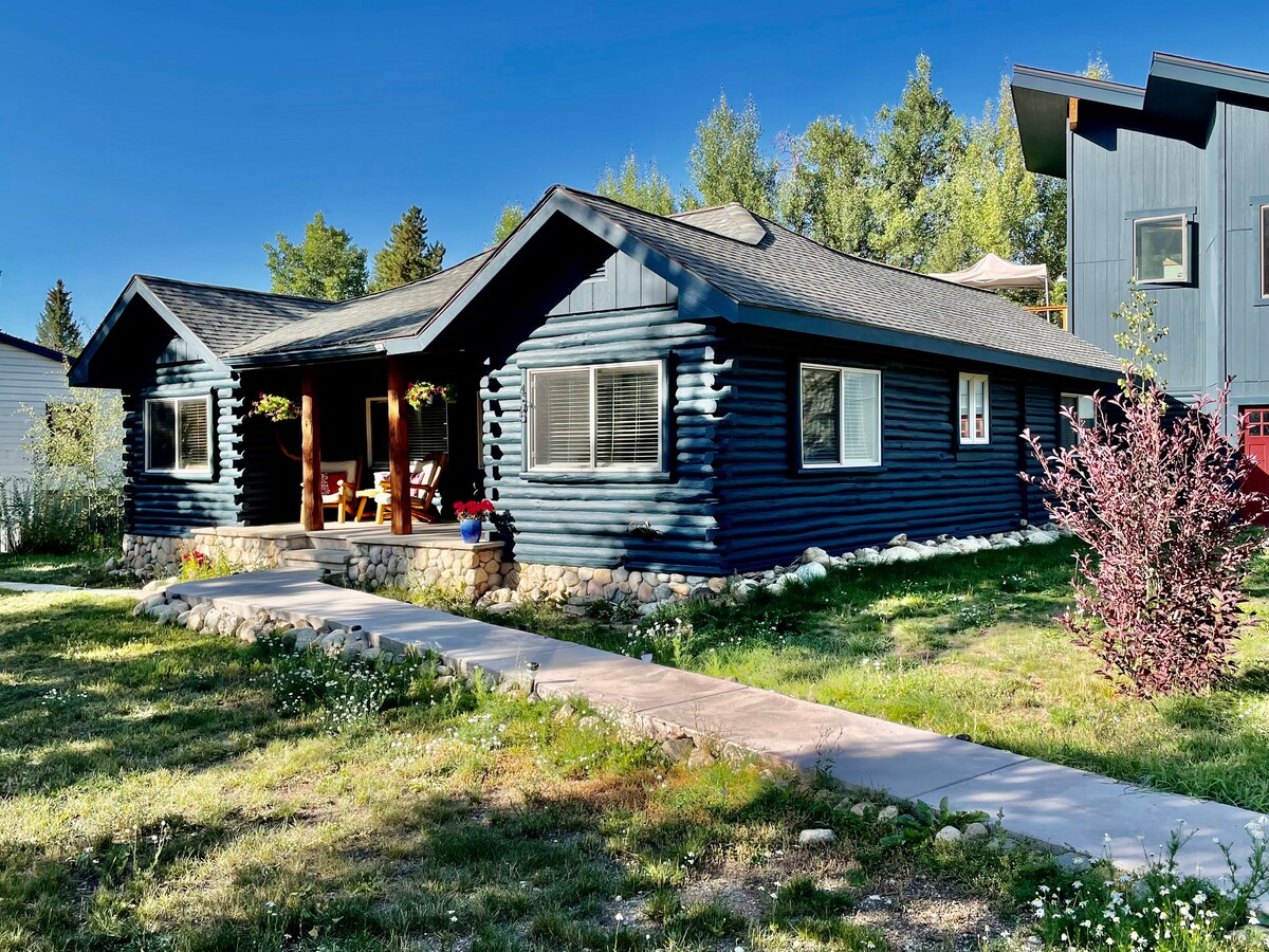 A charming log cabin with a dark blue exterior is framed by lush greenery. A pathway leads to the entrance adorned with flower planters, while large windows provide ample natural light to the interior. The welcoming porch features seating, perfect for enjoying the outdoors.