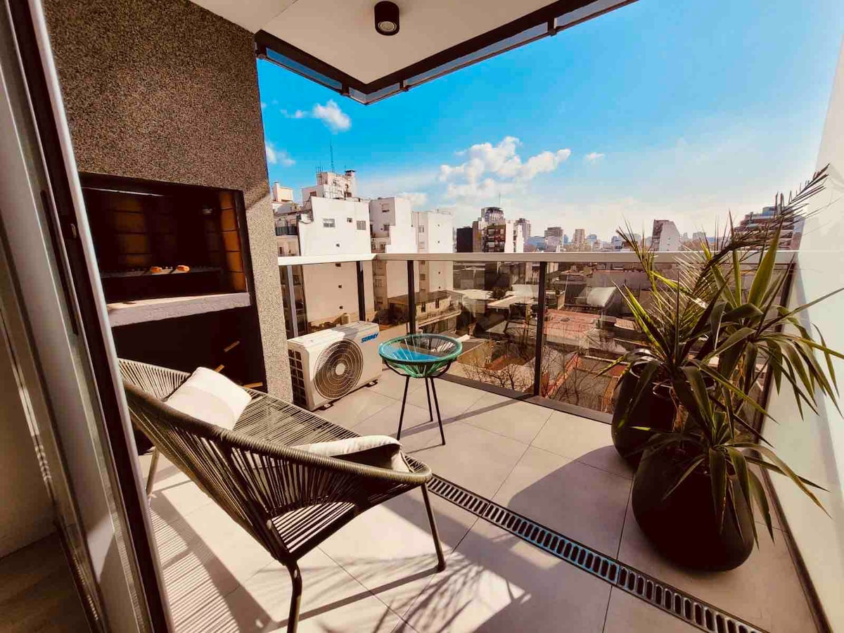 A balcony space features a modern grill and a pair of potted plants. A stylish chair and a small table provide seating, surrounded by a clear view of urban buildings under a bright blue sky. Sunlight creates an inviting atmosphere across the floor.