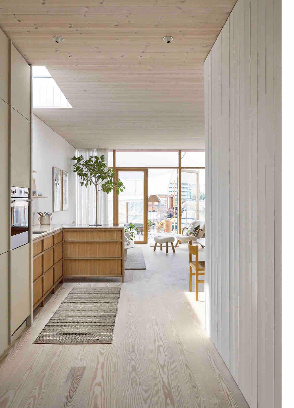 A bright interior space is seen, featuring light wood flooring and soft white walls. Natural light enters through large windows at the far end, illuminating a cozy seating area. A wooden sideboard and plants add to the harmonious ambiance.