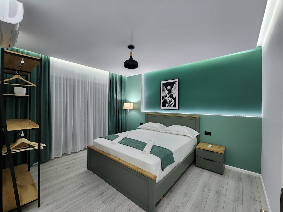 A modern sleeping area features a spacious bed with a light-colored headboard and green accents. A wooden bedside table and a clothing rack are present. Soft lighting is provided by a standing lamp, while sheer curtains allow natural light to filter in through the window.
