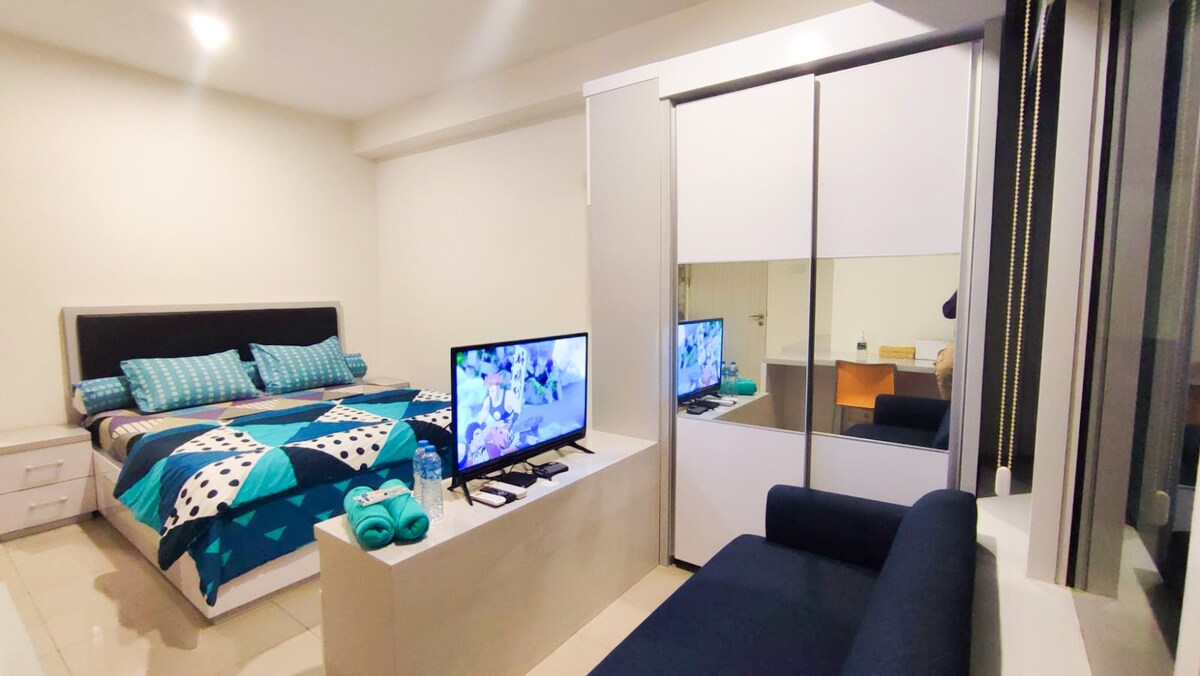 A well-appointed studio room features a spacious bed with patterned bedding and coordinating accent pillows. A television is positioned on a light-colored console. A modern cabinet stands against the wall, while a small seating area is provided by a dark couch, enhancing the inviting layout.