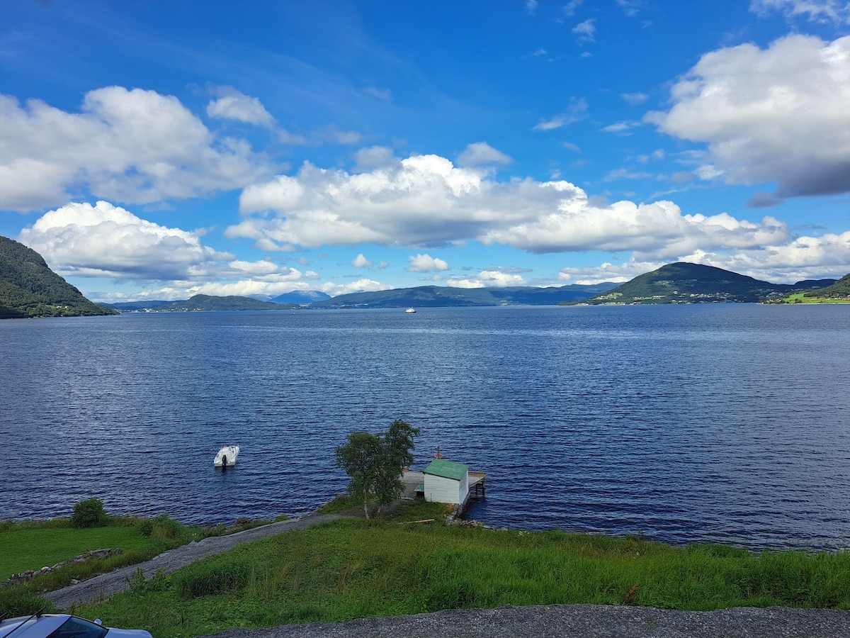 A serene view of Hjørundfjorden is displayed, featuring calm waters reflecting the sky's blue hues. A small boathouse is visible near the shore, surrounded by lush greenery and gentle slopes leading to the water's edge. Mountains frame the background, enhancing the natural scenery.