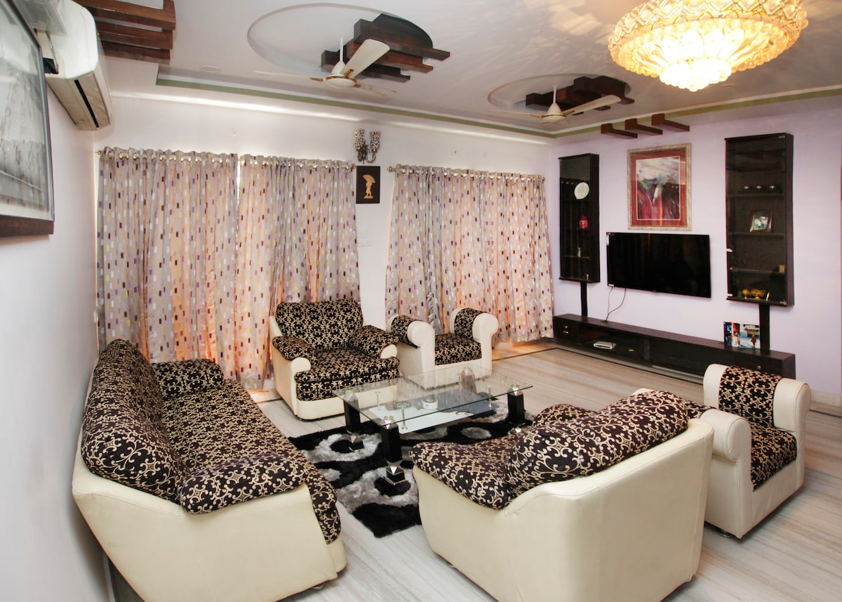 A spacious living area is furnished with matching patterned sofas and armchairs arranged around a glass coffee table. Light-colored curtains with polka dots frame the windows. A wall-mounted TV is visible, and a decorative ceiling light adds to the room's ambiance.