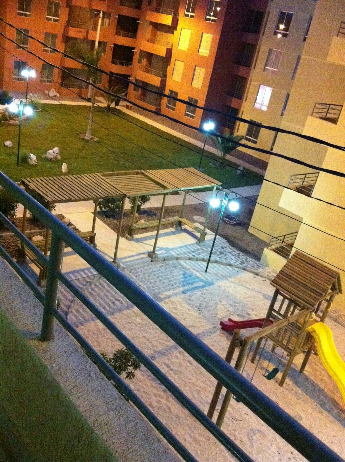 The image captures a playground area within a well-maintained community, featuring a wooden play structure with slides and climbing elements. Surrounding the play area is green grass, illuminated by nearby lights, along with a glimpse of building facades in the background.
