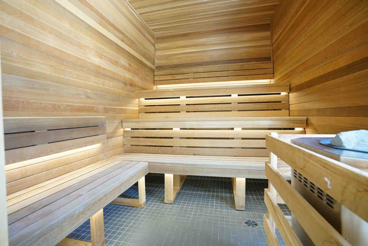 A wooden sauna features warm, natural wood paneling and built-in benches providing seating for several guests. Soft lighting accents the space, enhancing the tranquil atmosphere. A stone heater is positioned in the corner, complementing the overall design for relaxation.