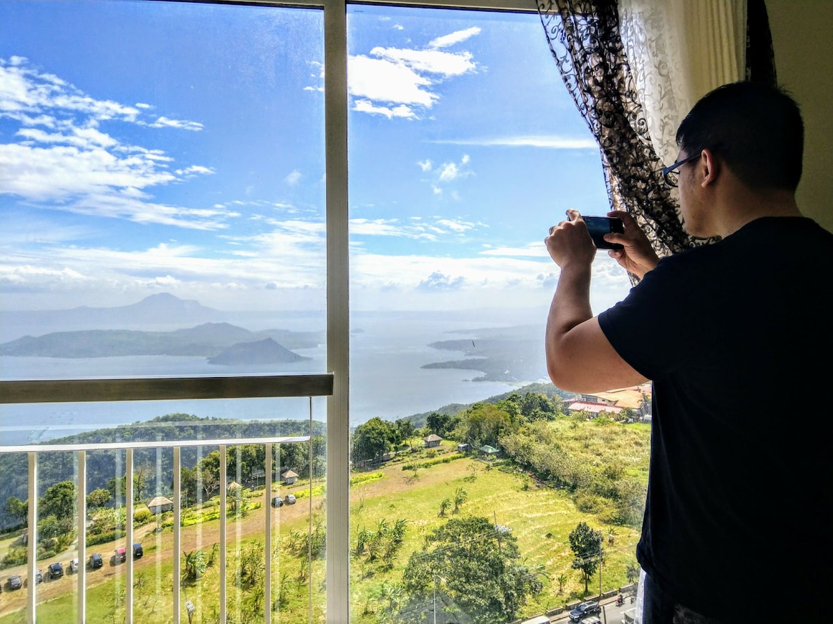A person is captured standing by a large window, taking a photo of the expansive view of the Taal Volcano and surrounding lake. Lush greenery is visible in the foreground, with distant mountains framed against a clear sky.