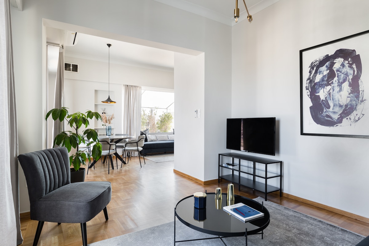 A bright living room features a comfortable gray armchair and a round coffee table. A flat-screen TV is positioned on a sleek black stand. Natural light floods the space through large windows, connecting it to a dining area with a stylish table and modern chairs.
