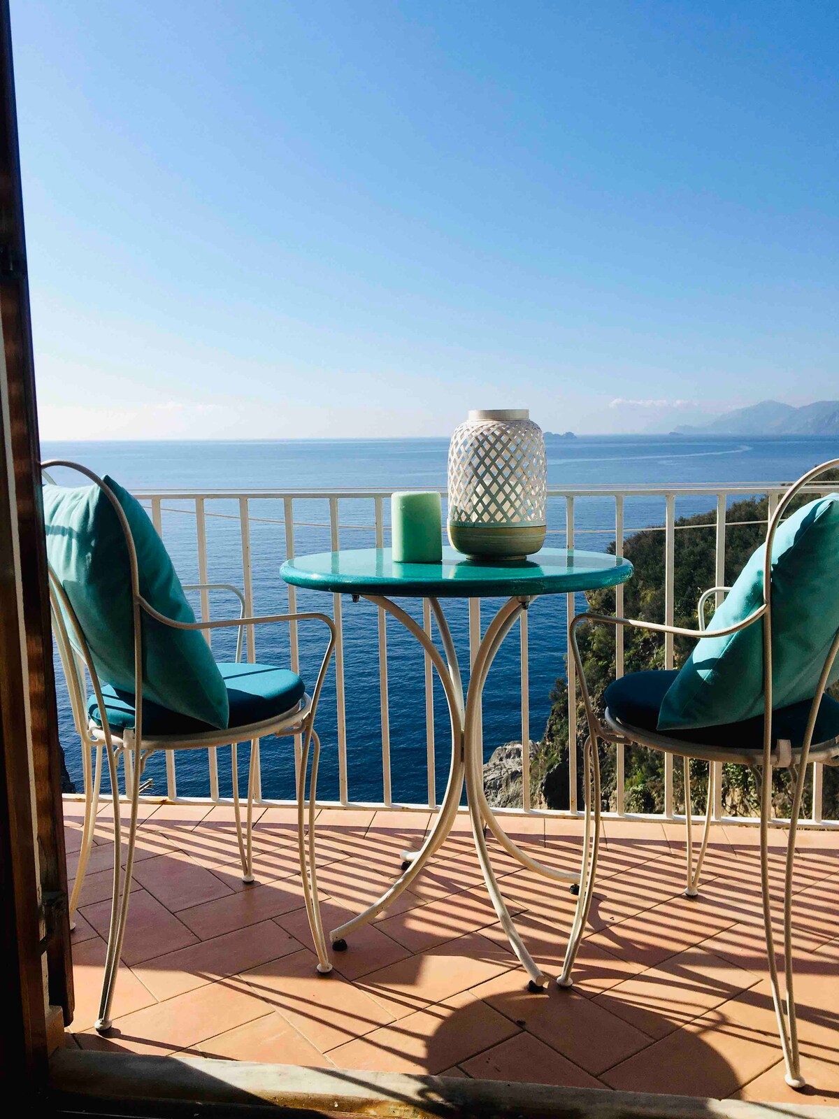 A small terrace is featured with a round table and two chairs, surrounded by scenic views of the emerald sea. A lantern and green candle offer subtle decor, while the backdrop showcases the horizon and rugged coastal landscape under a clear blue sky.
