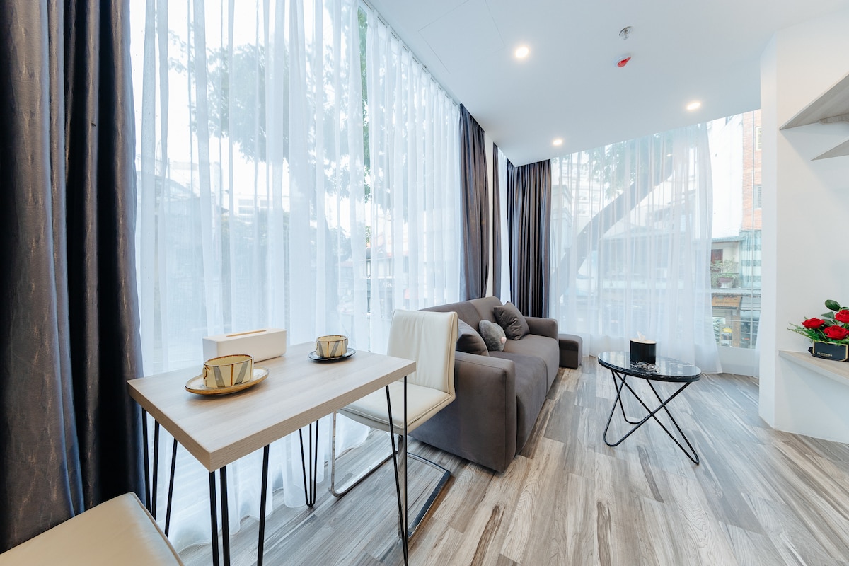 A light-filled living area is presented with large windows draped in sheer curtains, allowing natural light to filter in. A comfortable gray sofa complements a modern coffee table, while a dining table with two place settings enhances the space's functionality.