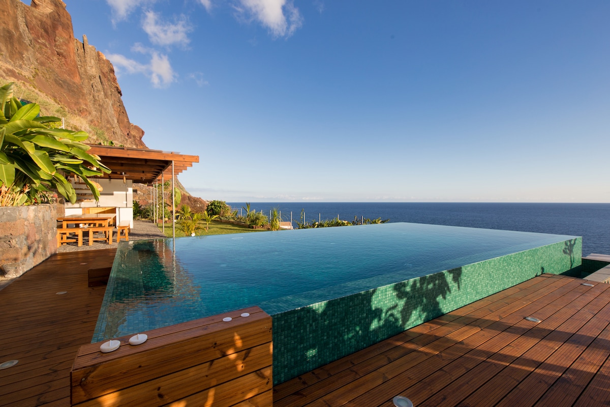 An infinity pool stretches over a wooden deck, with views of the expansive Atlantic Ocean and dramatic cliffs. Sunlight reflects off the water's surface, enhancing the serene atmosphere. Surrounding greenery complements the natural setting, inviting relaxation and contemplation.