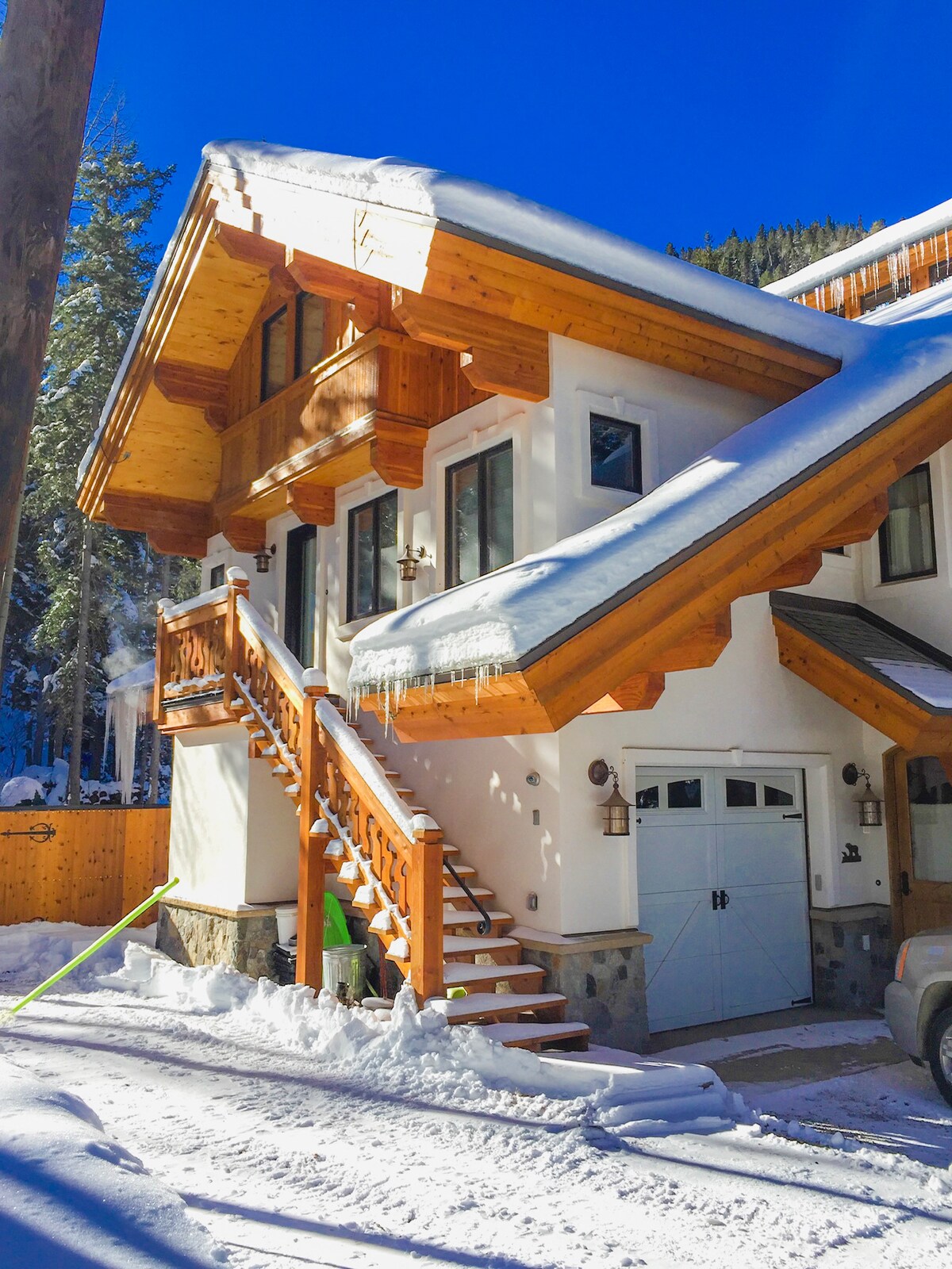 A charming exterior of a Bavarian-style apartment is displayed, featuring wood accents and a sloped roof. Snow blankets the surrounding area, and a staircase leads to the entrance. Large windows and warm wood details contribute to the inviting appearance of the structure.