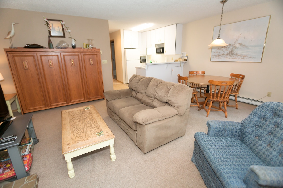 An inviting living area features a cozy beige sofa and a patterned blue chair, complemented by a wooden coffee table. A charming dining set with four wooden chairs and a round table is visible in the background, along with a well-equipped kitchen area.