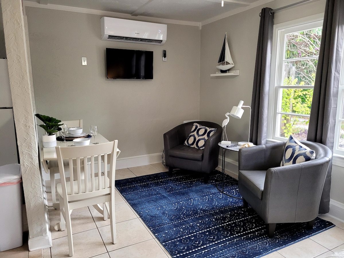A cozy living area features two grey armchairs with patterned cushions, accompanied by a small round table. A wall-mounted television and nautical décor are visible. Natural light filters through the large window, enhancing the neutral tones of the space.