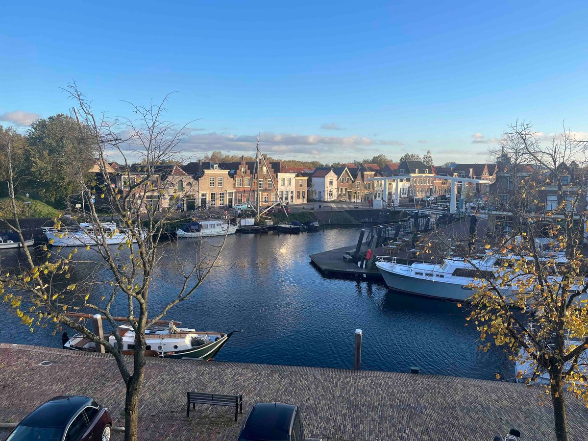 A scenic view of a marina is presented, showcasing several boats moored along a calm waterway. Colorful buildings line the waterfront, and trees with autumn leaves frame the scene. A bright sky with soft clouds adds to the tranquil atmosphere.