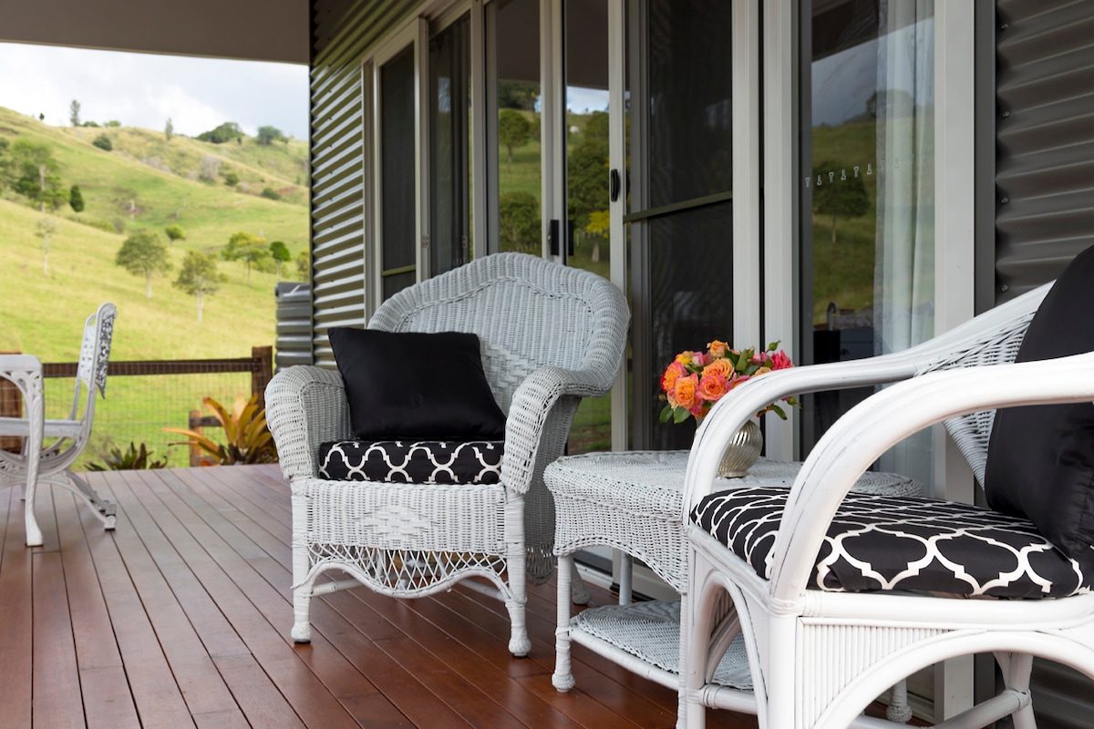 A porch area features white wicker chairs with dark cushions, arranged invitingly on polished wooden flooring. A small table holds a vase of colorful flowers. The background showcases rolling hills, emphasizing the tranquil rural setting.
