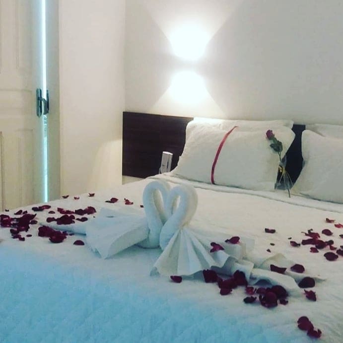 A neatly arranged bed is displayed in a softly lit room. Elegant towel swans are crafted at the center of the bed, surrounded by scattered rose petals. A simple decoration of a rose is seen on the headboard, enhancing the room's serene atmosphere.