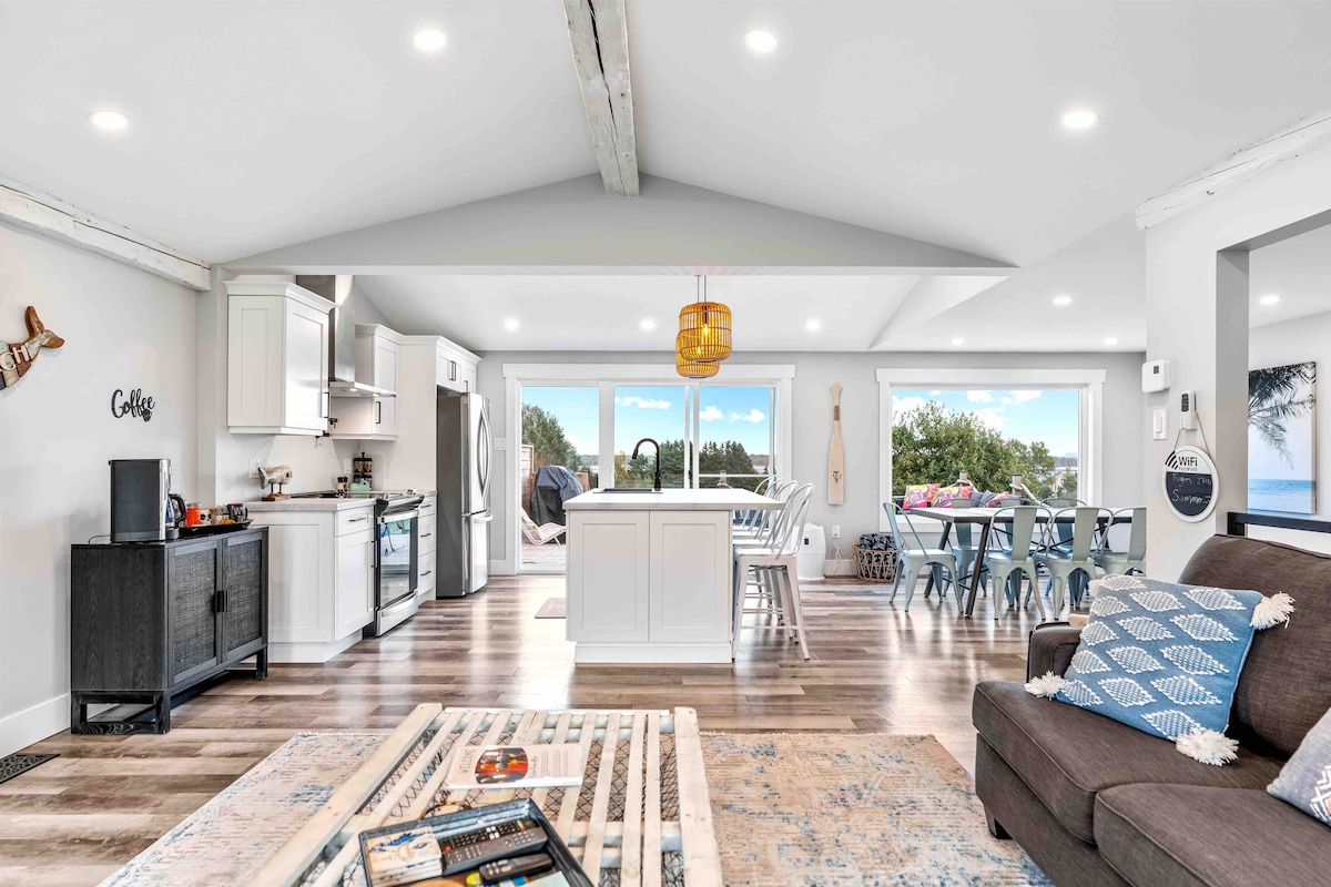 An open-concept kitchen and living area is presented, featuring modern white cabinetry and stainless steel appliances. A large island with seating is visible, along with a comfortable seating arrangement. Natural light fills the space through expansive glass sliding doors, highlighting the scenic outdoor view.